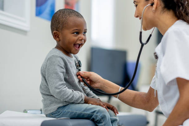 A young boy of African decent, sits up on an exam table as a female Paediatrician preforms a check-up on him.  The boy is dressed casually and smiling as the doctor listens to his heart.